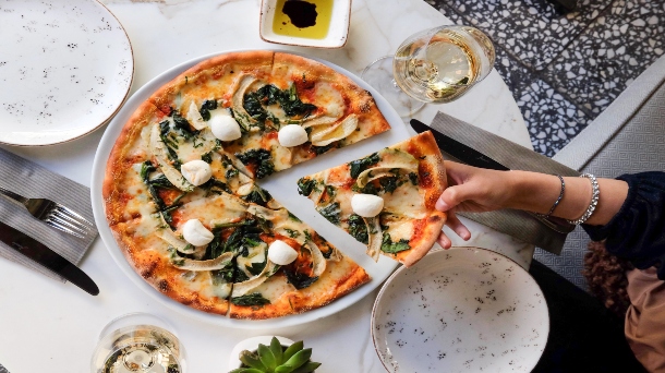 Na bílém kulatém talíři je pizza nakrájená na 6 dílků. Ruka ženy si bere jeden dílek pizzy z talíře .